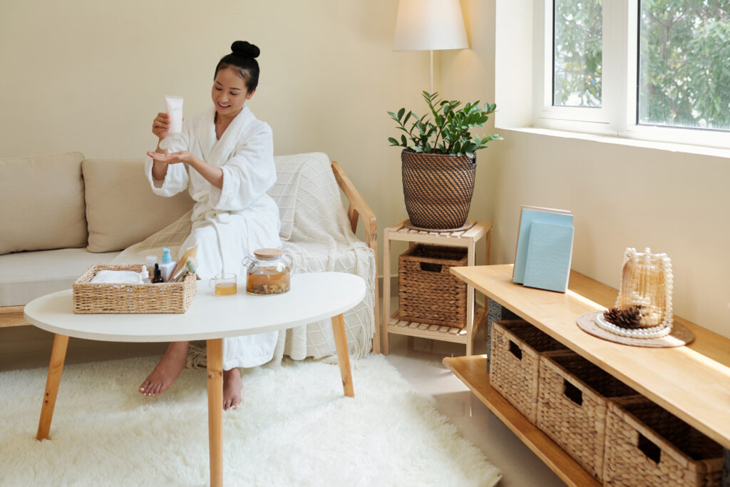 Positive young Asian woman drinking herbal tea and applying moisturizing lotion after taking bath