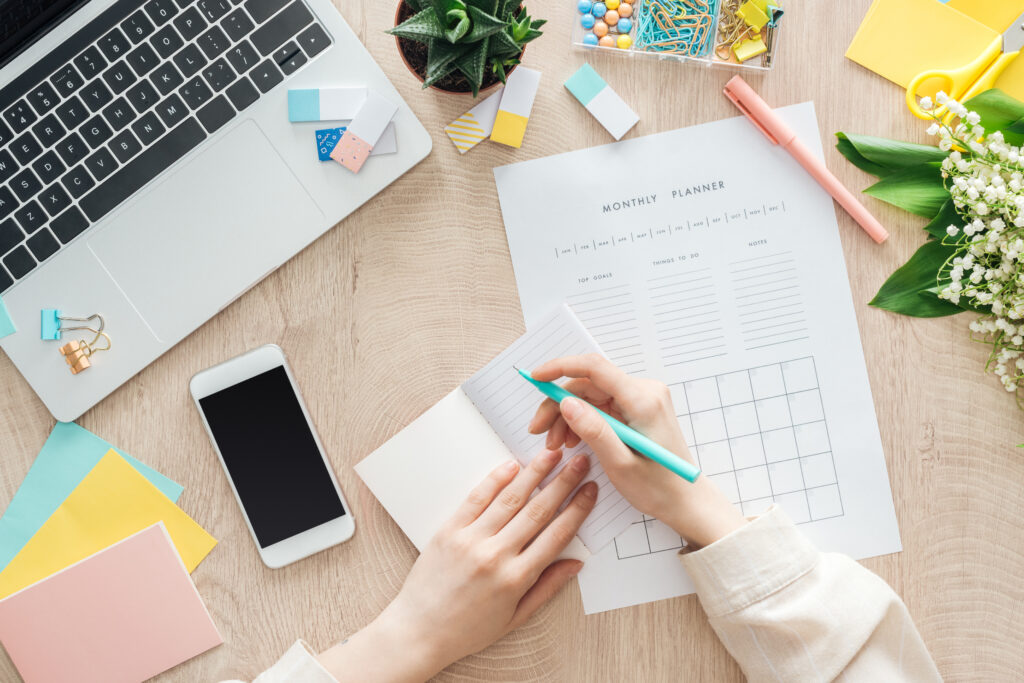 cropped view of woman sitting behind wooden table with smartphone, laptop and stationery, writing in