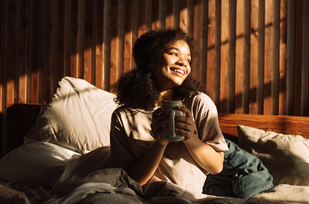 Charming mixed-race teenager is laughing and holding cup of coffee while sitting in bed. Pretty African girl is enjoying sunny morning at home. Cute student is drinking tea and looking out window.