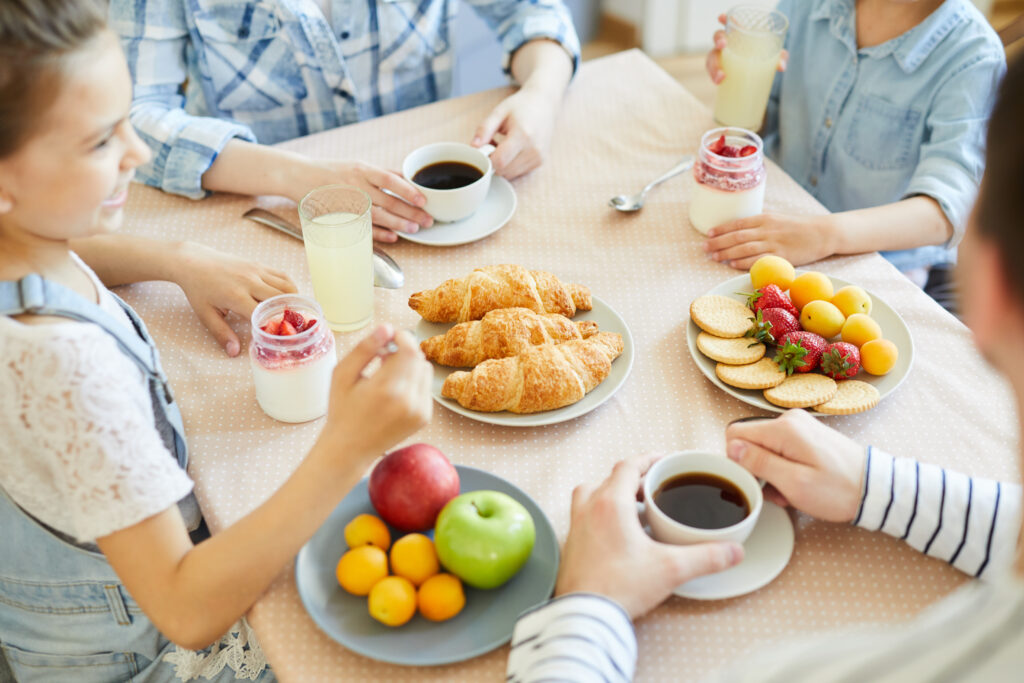 Parents having coffee and little siblings eating strawberry dessert by table served with fruits, croissants and biscuits