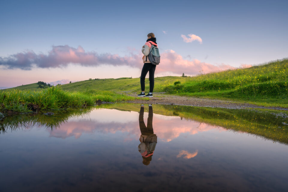 Woman with backpack reflected in lake at sunset in summer. Velika Planina, Slovenia. Beautiful girl on the shore of pond is looking on green meadows and blue sky with pink clouds at dusk. Hiking